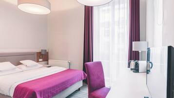 A hotel room with a bed, a chair, a desk with a lamp, a mirror, and a window with curtains.