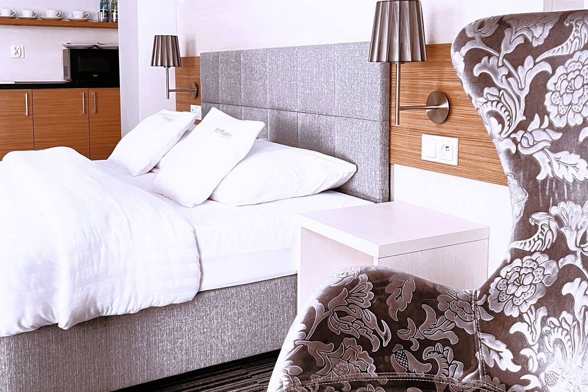 A modern hotel room with a bed, bedside table, lamp, chair, and a small kitchenette.