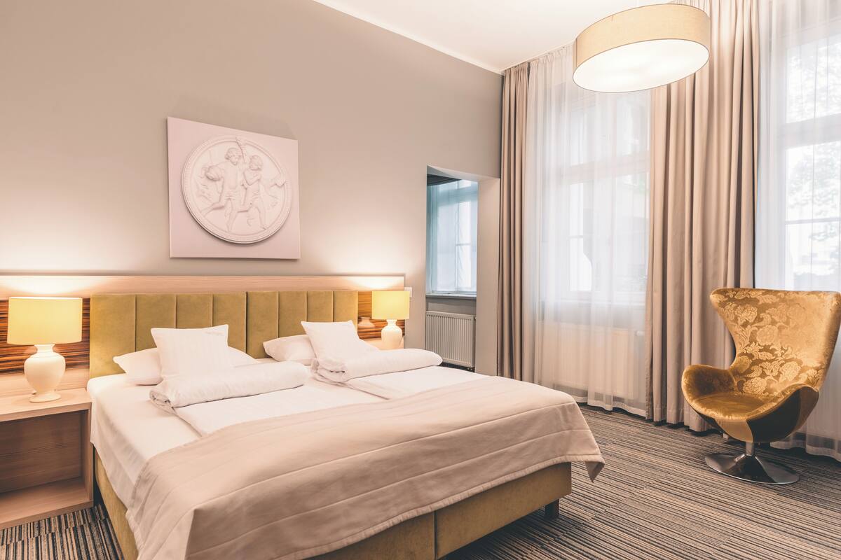A hotel room with a large bed, a nightstand with a lamp, a chair, and a window with curtains.