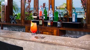 A bar with a view of the ocean, wooden accents, and various bottles and glasses. - Xbalanque (Roatan)