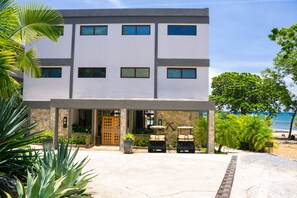 A modern two-story building with a covered entrance, outdoor seating, and a view of the beach.