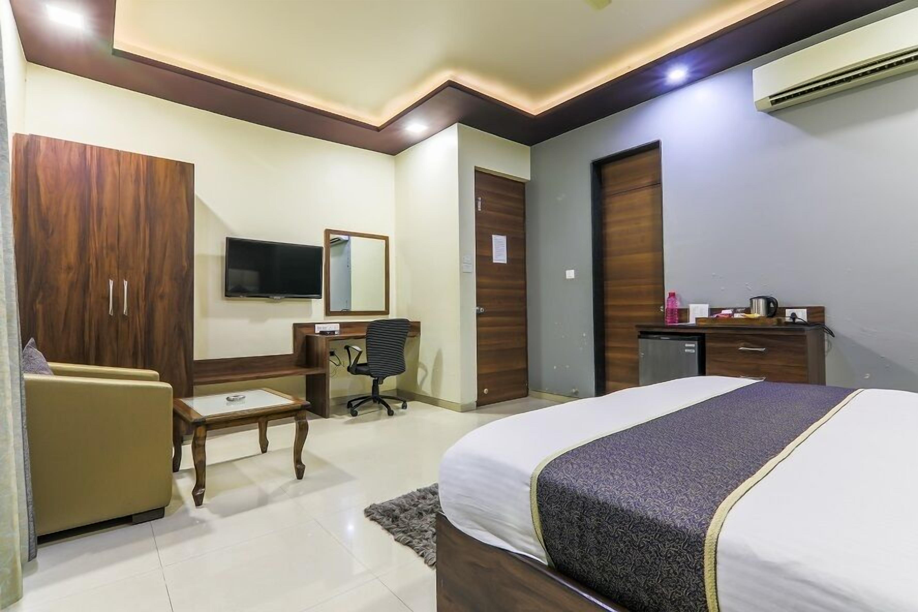 A hotel room with a bed, a desk, a chair, a wardrobe, a television, and an air conditioning unit.