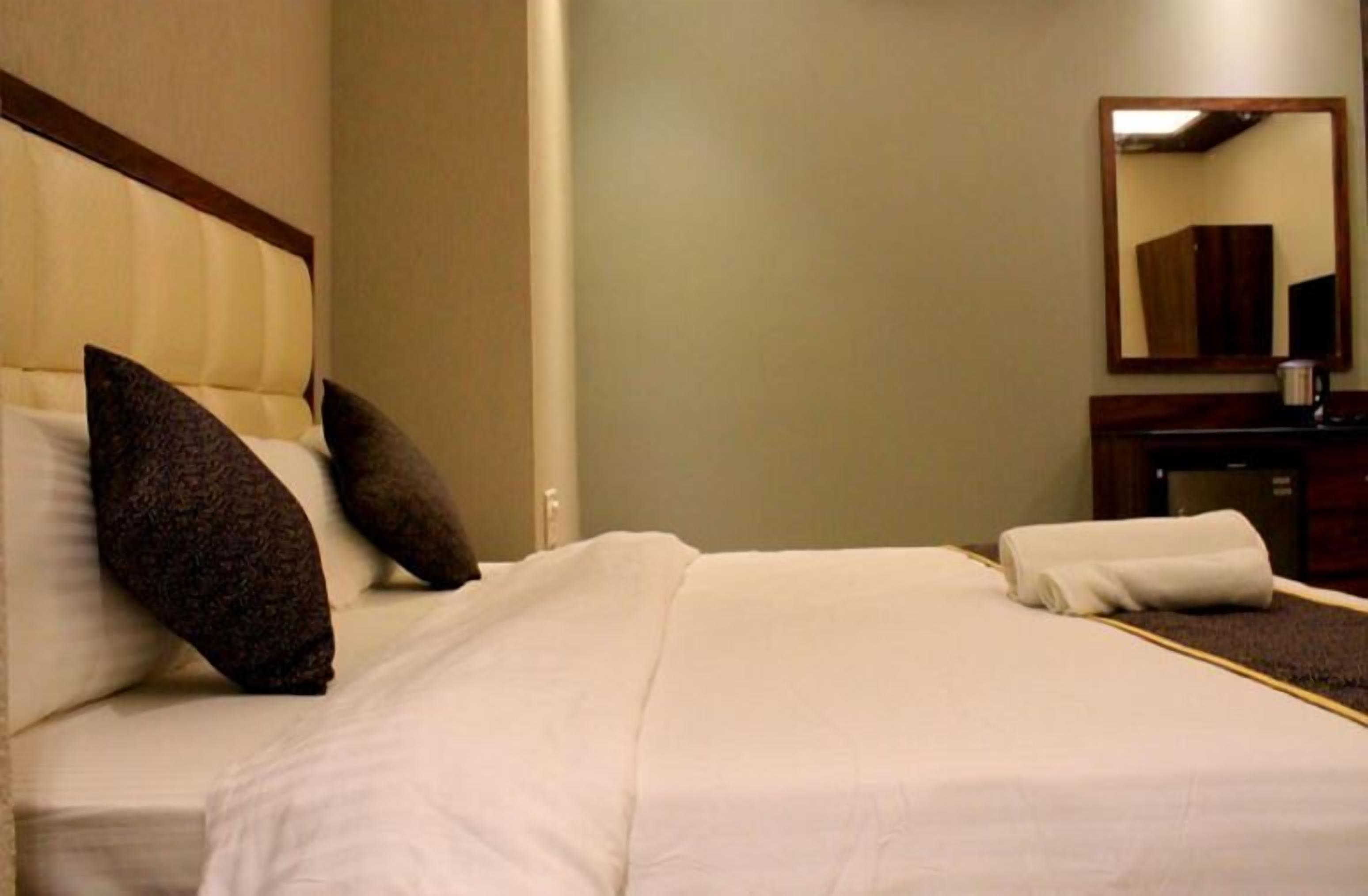 A hotel room with a bed, pillows, a bedside table, a mirror, and a small cabinet.