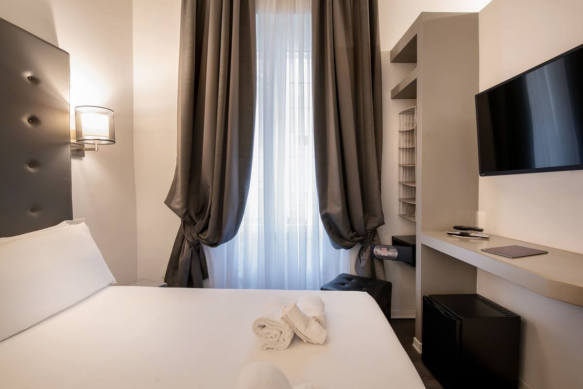 A hotel room with a bed, a TV, a desk, and a window with curtains.