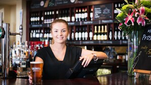 A bar with a woman holding a glass, a cash register, and a display of wine bottles in the background. - Rose & Crown (Plymouth)