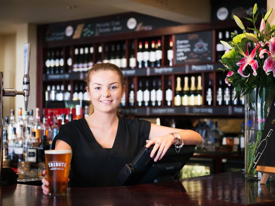 A bar with a woman holding a glass, a cash register, and a display of wine bottles in the background.