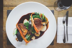 A white plate with a dish of food, a fork and knife on a napkin, and a glass of dark liquid on a wooden table. - Rose & Crown (Plymouth)