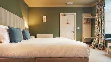 Una cama bien hecha con ropa de cama blanca y azul, un radiador blanco, una puerta con un abrigo colgado y una ventana con cortinas estampadas.