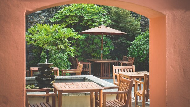An outdoor patio area with wooden tables and chairs, a stone fountain, and lush greenery. - Rose & Crown (Plymouth)