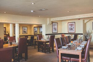 A restaurant with multiple dining tables and chairs, framed pictures on the walls, and a carpeted floor.