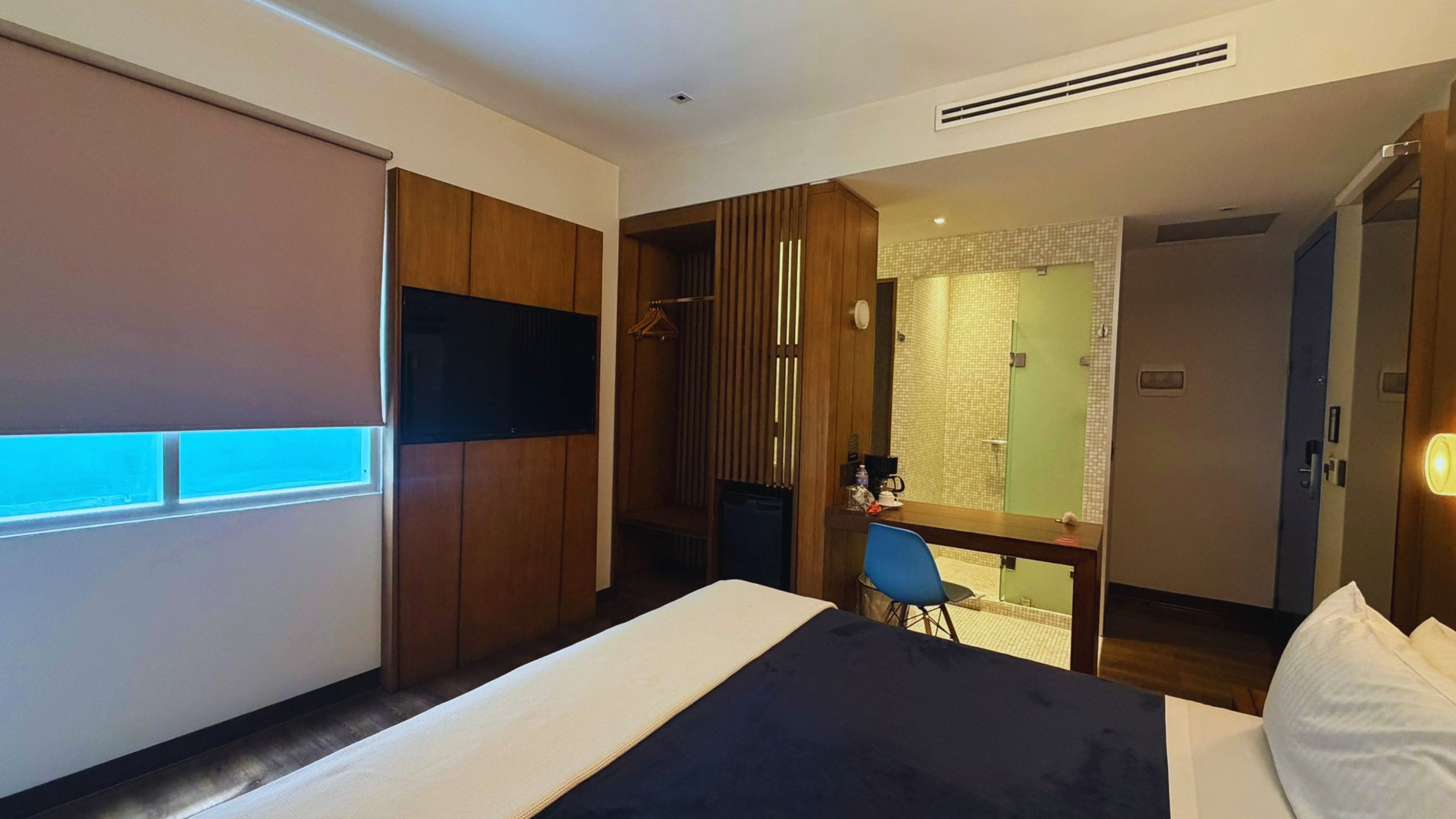Habitación de hotel con una cama, un escritorio, una silla, un televisor y un baño visible a través de una puerta abierta.