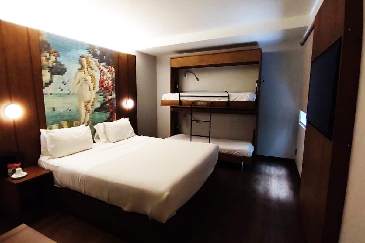 A hotel room with a large bed, a bunk bed, a wall-mounted TV, and a painting on the wall.