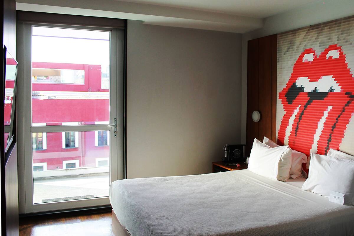 A hotel room with a bed, a window, and a wall with a red and white pattern.