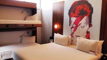 A hotel room with a bunk bed, a single bed, a wall-mounted artwork, and a bedside table with a lamp.