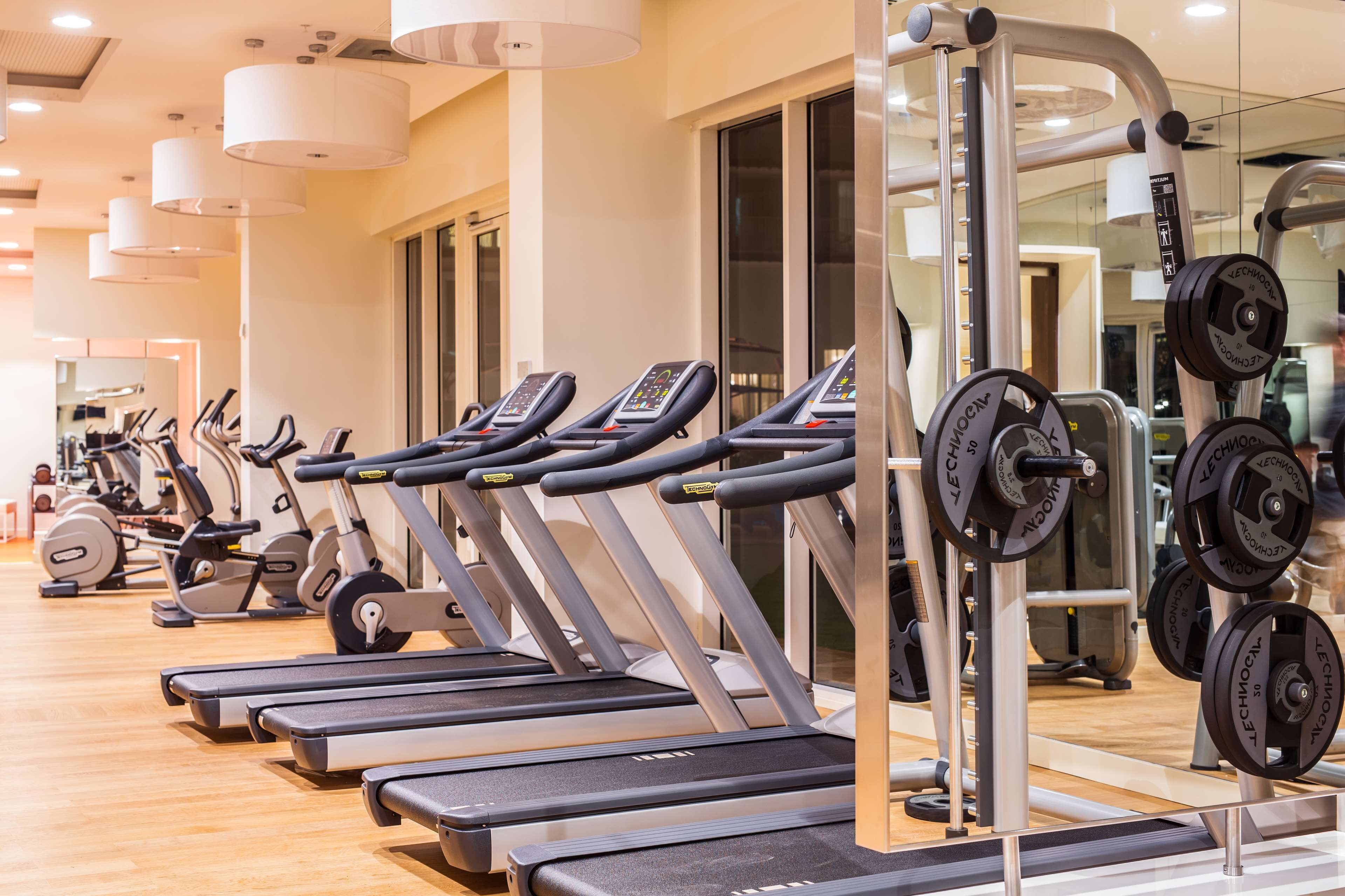 En række løbebånd i et fitnesscenter med styrketræningsudstyr og spejle på væggen.