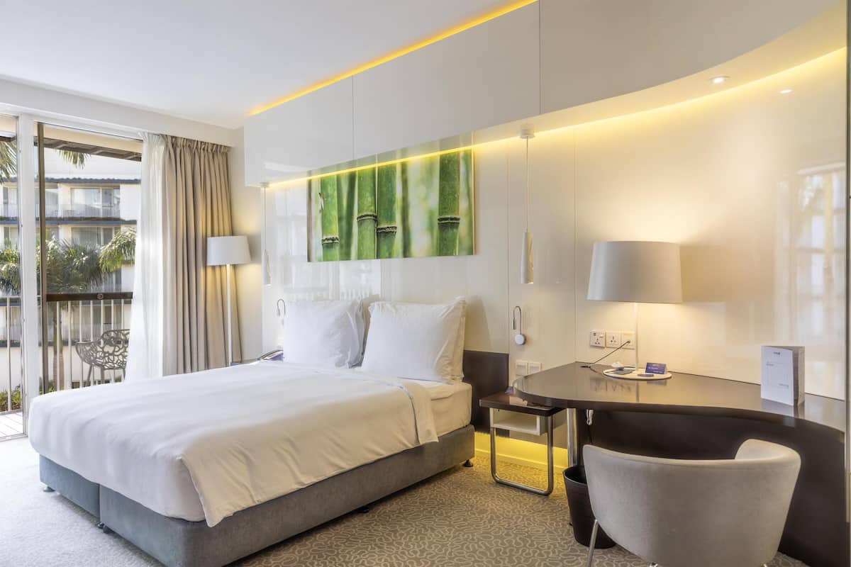A hotel room with a bed, a desk, a chair, a large window, and a view of the outside.