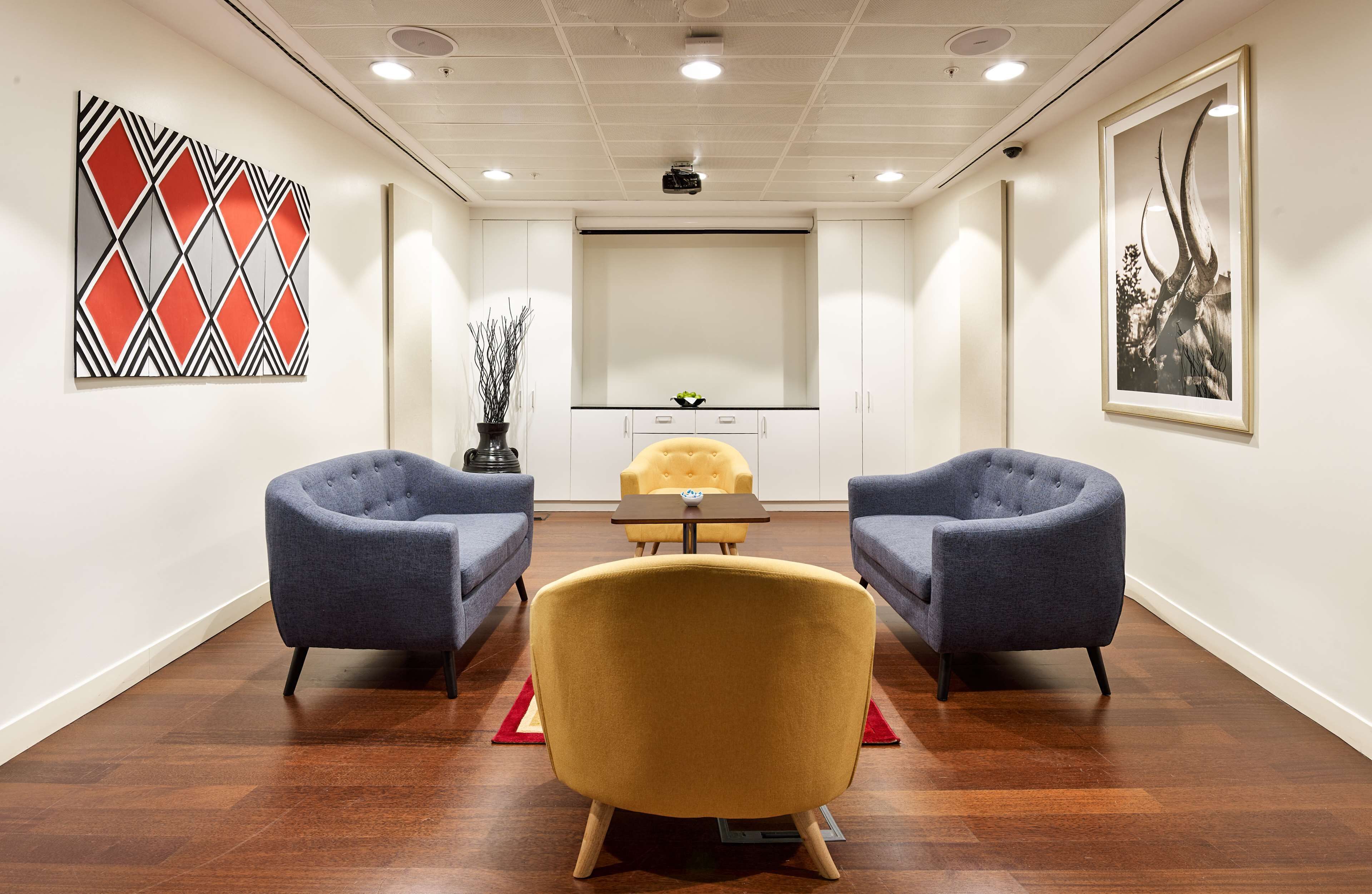 A conference room with two blue armchairs, a yellow chair, a small table, and a projector screen.