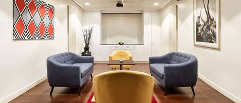 Une salle de conférence équipée de deux fauteuils bleus, d’une chaise jaune, d’une petite table et d’un écran de projection.