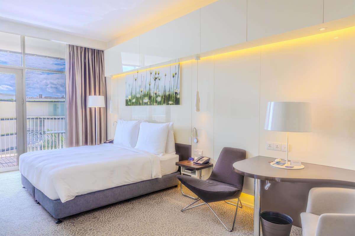 A hotel room with a bed, a desk, a chair, a painting, and a view of the outdoors.