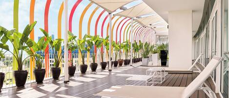 A sunlit outdoor deck with lounge chairs and potted plants, featuring a curved ceiling with colorful vertical stripes.