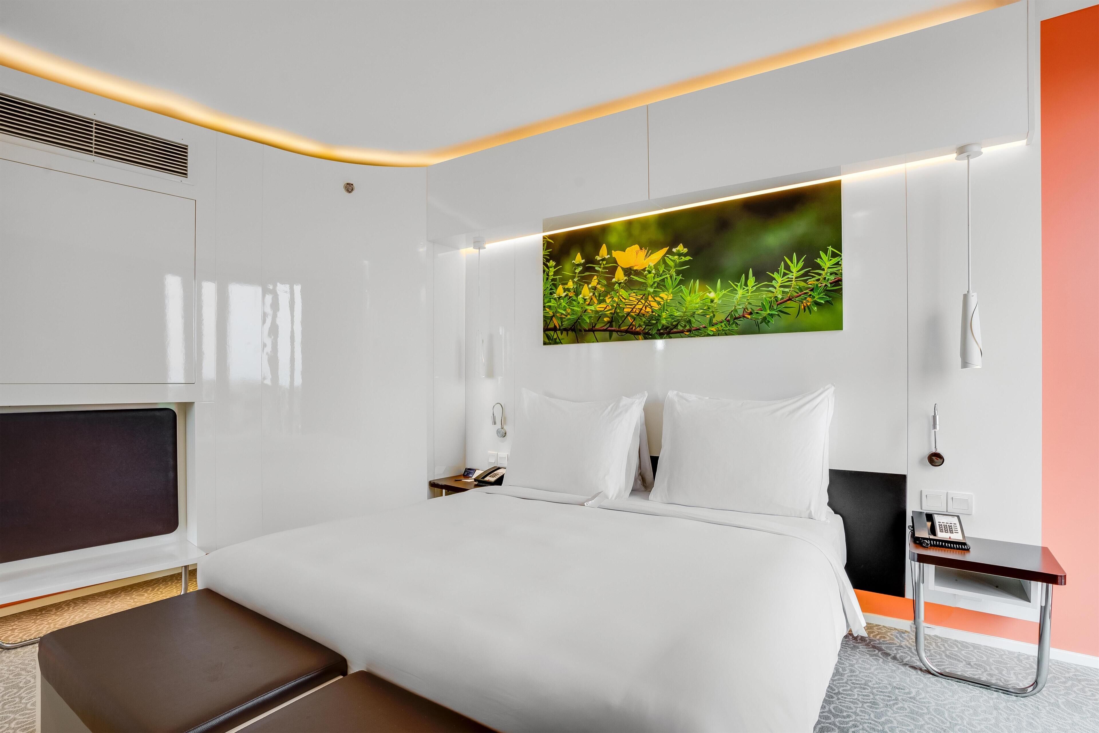 Una habitación de hotel moderna con una cama grande, un televisor de pantalla plana, una mesita de noche con teléfono y una obra de arte colgada en la pared.