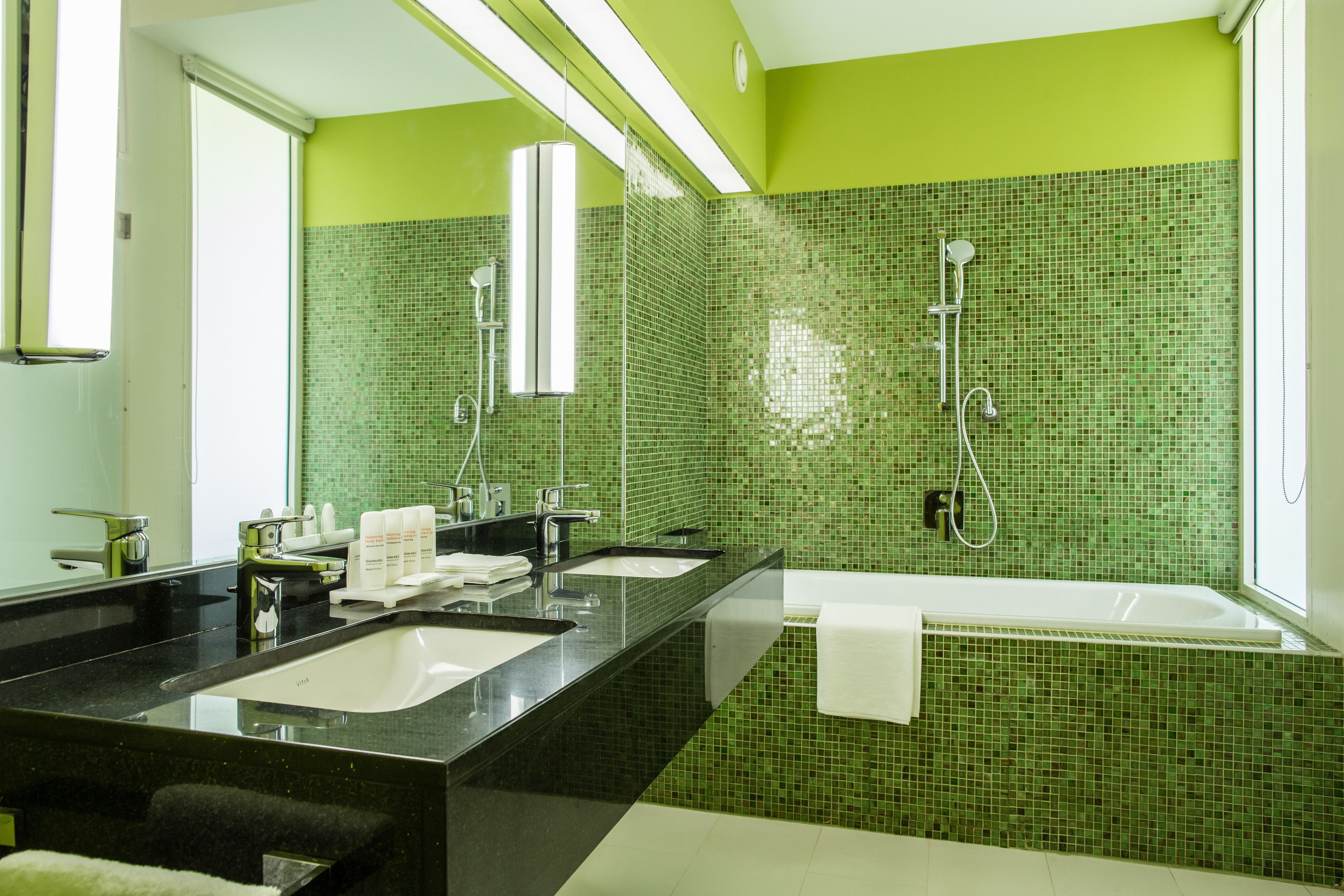 A modern bathroom with green mosaic tiles, a black countertop, a large mirror, and a bathtub.