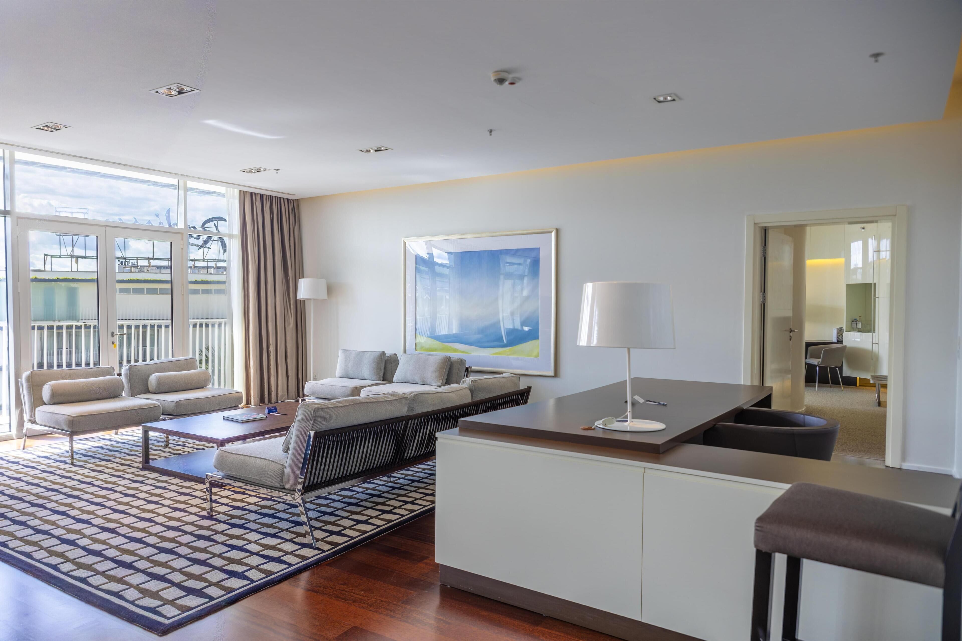 A modern hotel room with a sofa set, a dining table, a kitchen area, and large windows.