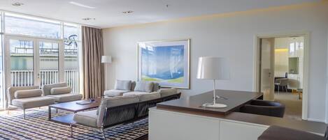 A modern hotel room with a sofa set, a dining table, a kitchen area, and large windows.