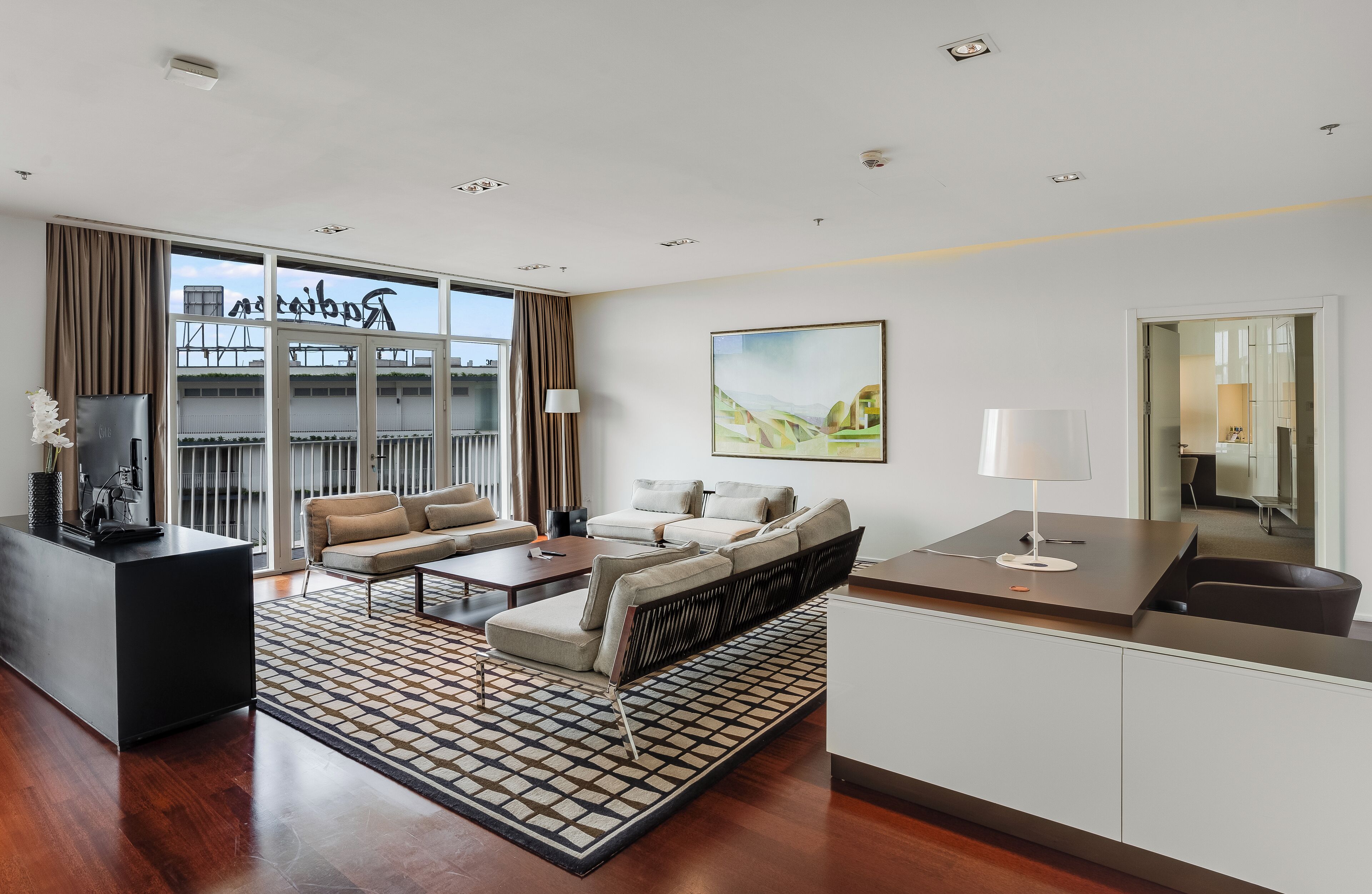 A modern hotel room with a sofa, coffee table, and a desk with a lamp. There is a large window with a view of buildings and a balcony.