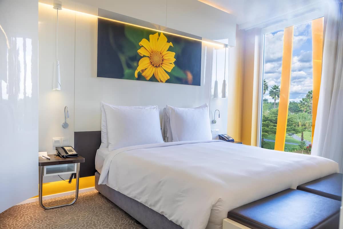 A hotel room with a large bed, a bedside table with a phone, a painting of a flower, and a window with a view of greenery.