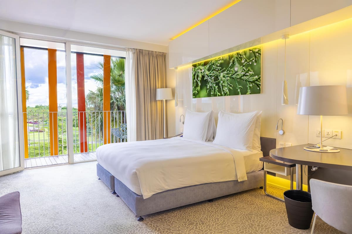 A hotel room with a bed, a desk, a chair, a large window with a view of greenery, and a balcony.