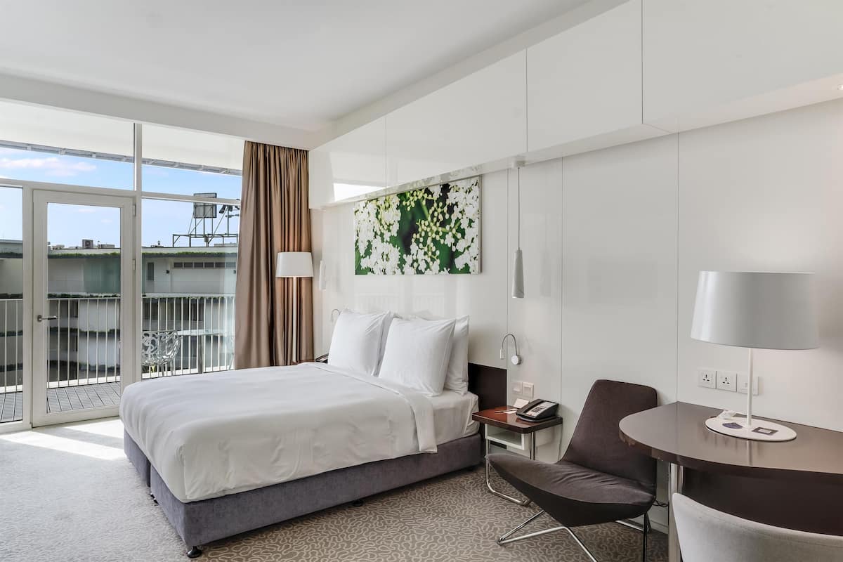 A hotel room with a bed, a desk, a chair, a painting, and a balcony with a view.