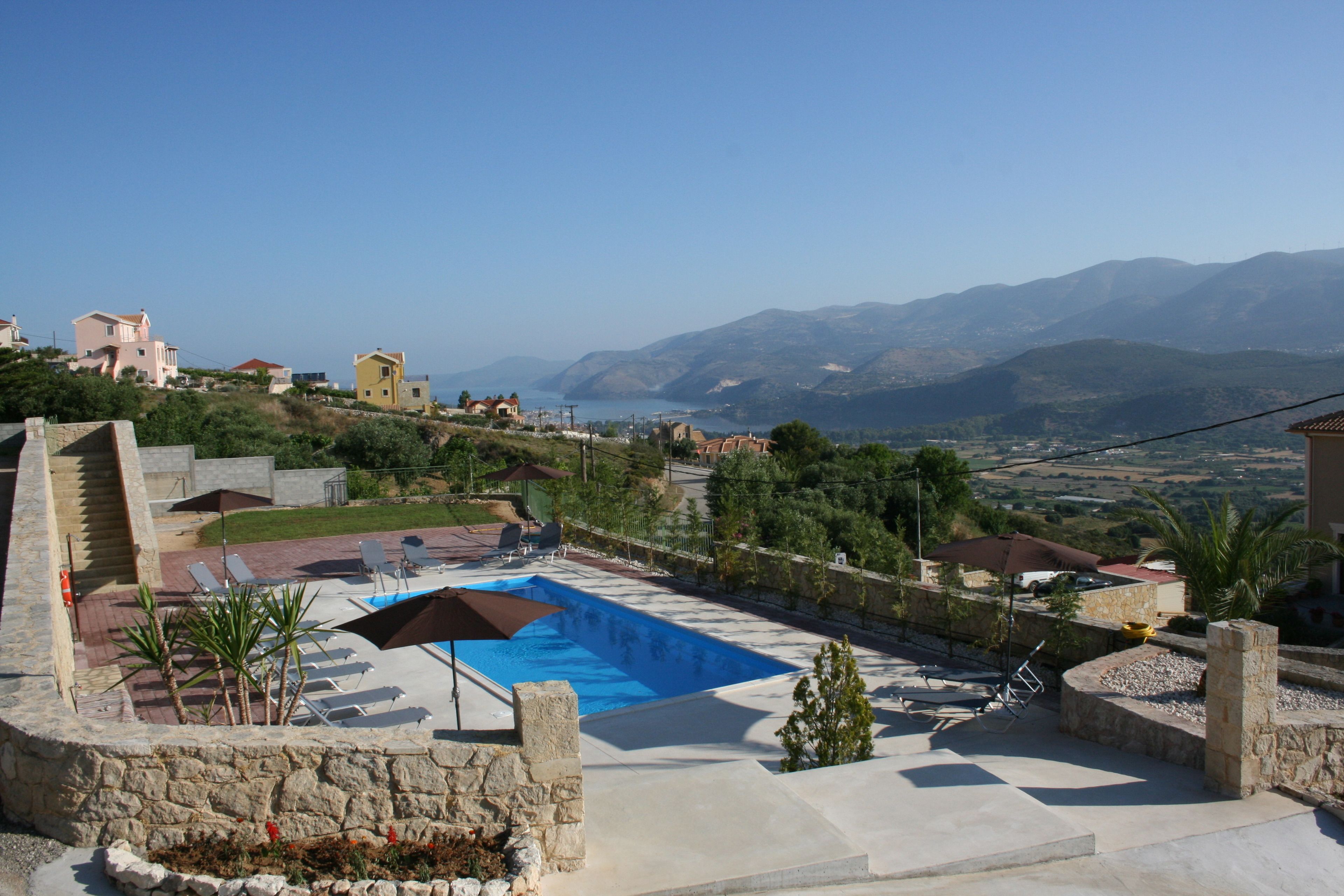 A swimming pool with lounge chairs and umbrellas, surrounded by stone walls and greenery, overlooking a mountainous landscape.