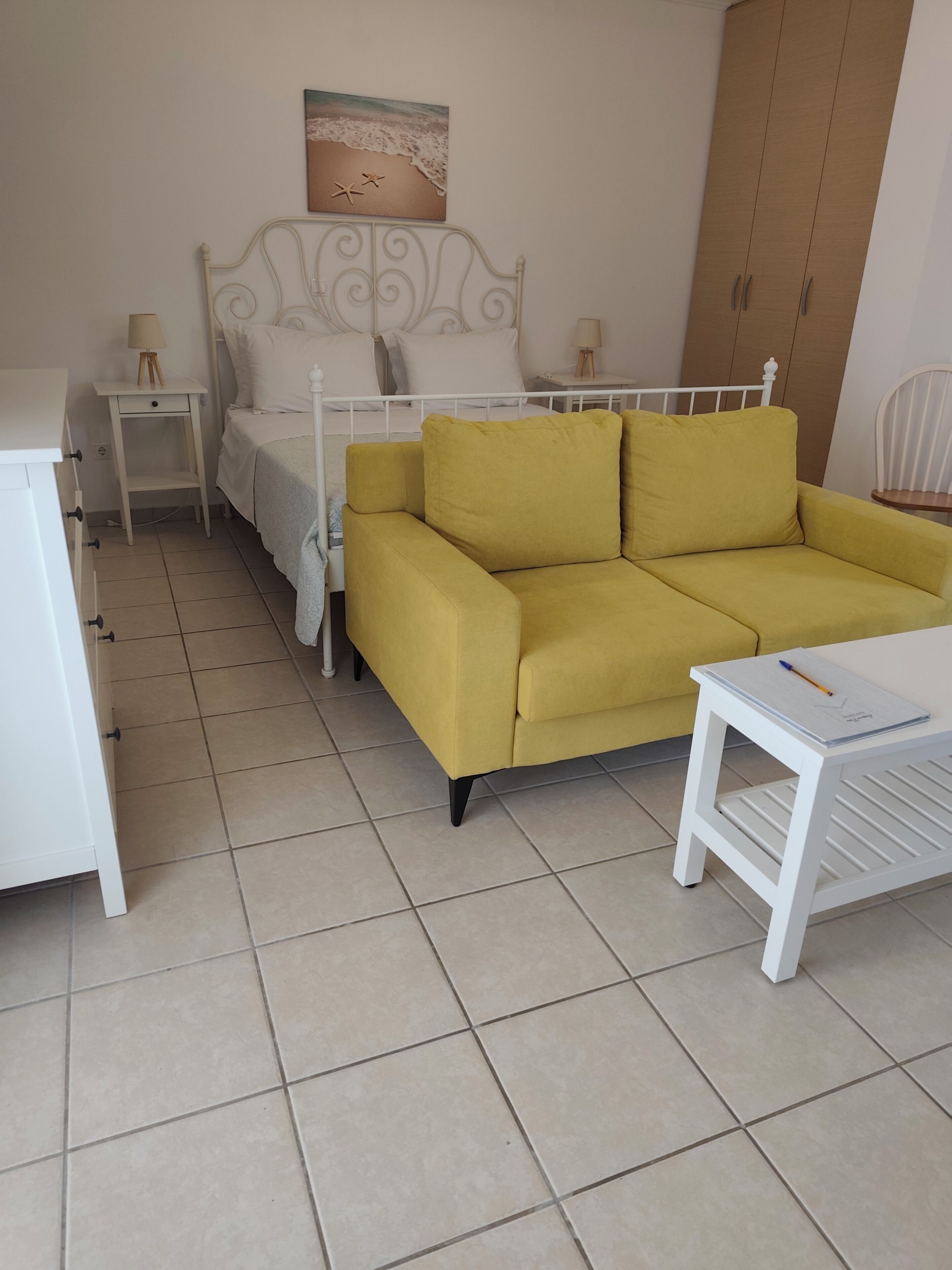 A room with a bed, a yellow sofa, a white nightstand, a white table, and a wooden wardrobe.