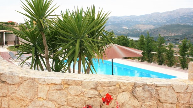 A stone wall with a pool, palm trees, and a view of mountains. - Lagoon View Apartments - Adults Only (Kefalonia)