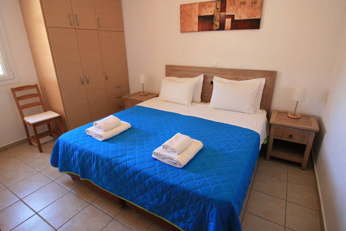 A neatly made bed with a blue bedspread and two white pillows, a wooden wardrobe, and a small bedside table with a lamp.