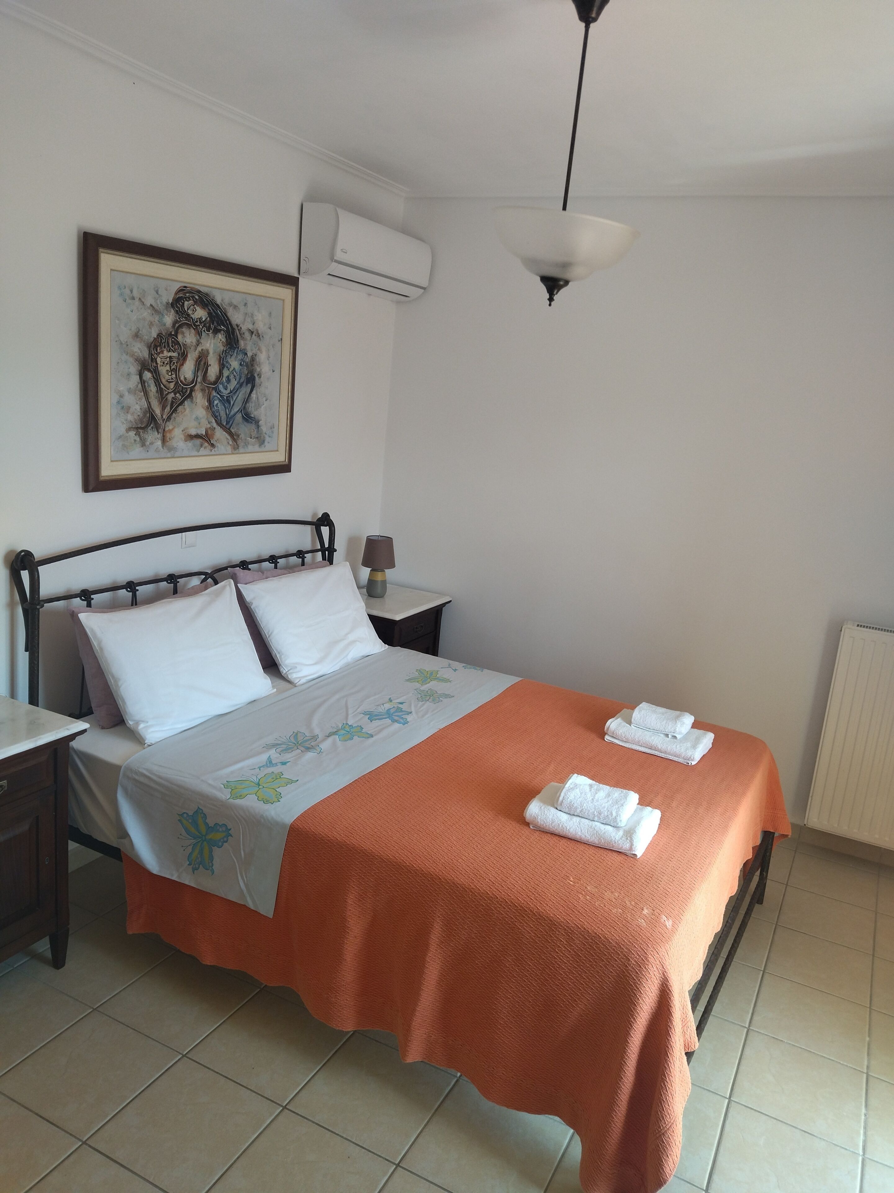 Una cama bien hecha con una colcha naranja y almohadas blancas, una lámpara de noche y una obra de arte enmarcada en la pared.