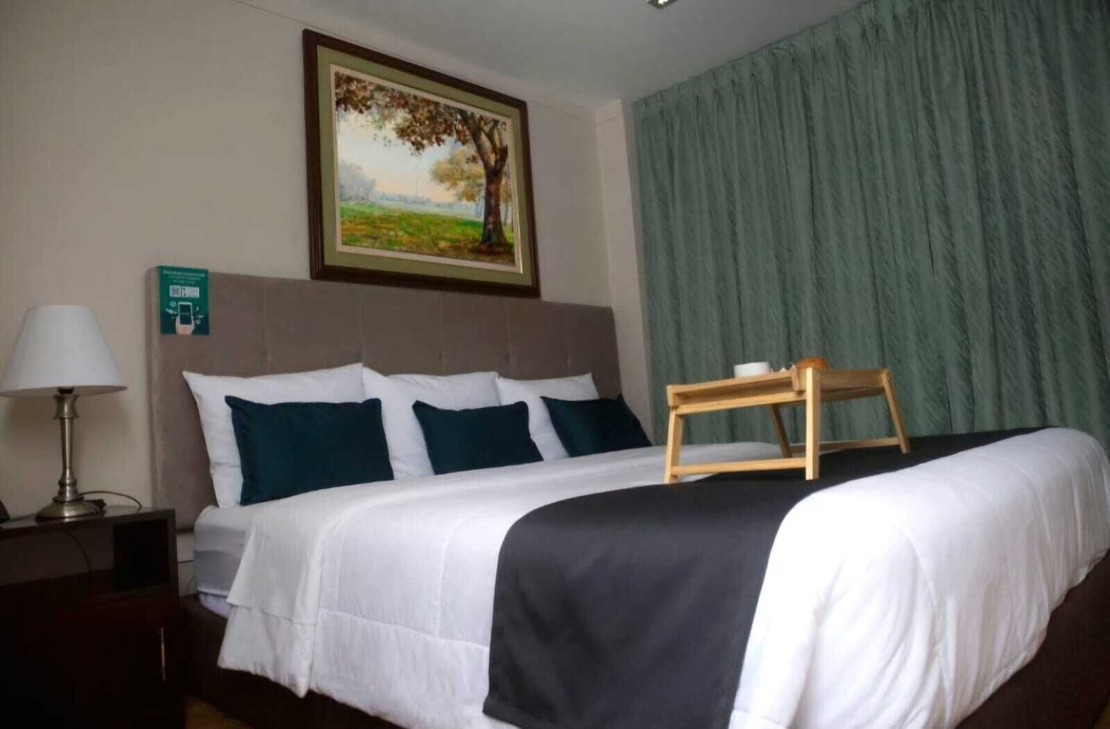 A hotel room with a large bed, a nightstand with a lamp, a painting of a tree, and a green curtain.