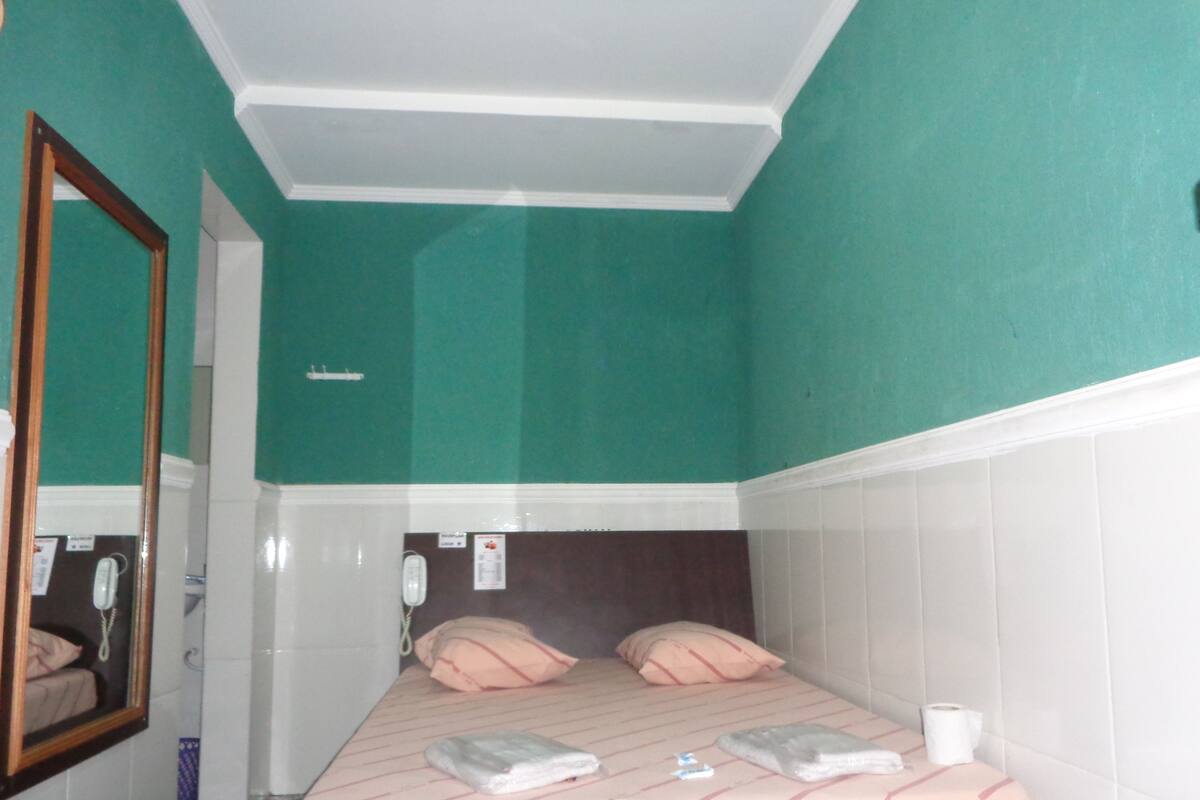 Ein Hotelzimmer mit türkisfarbenen Wänden, einem Bett mit pinkener Bettwäsche, einem Spiegel und einem Nachttisch mit einer Tasse.