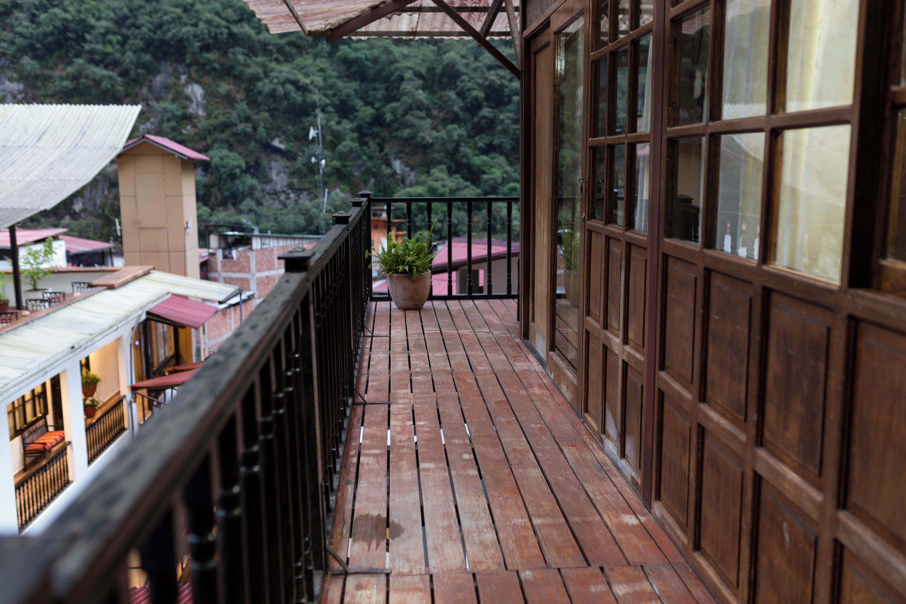 A wooden balcony with a black railing, potted plants, and a view of buildings and greenery in the background.