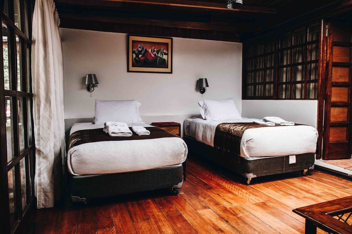 A room with two beds, wooden flooring, a painting on the wall, and a window with curtains.