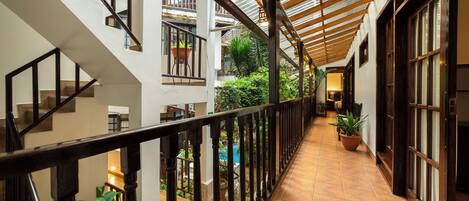 A covered corridor with a wooden railing, potted plants, and a view of a pool and greenery outside.