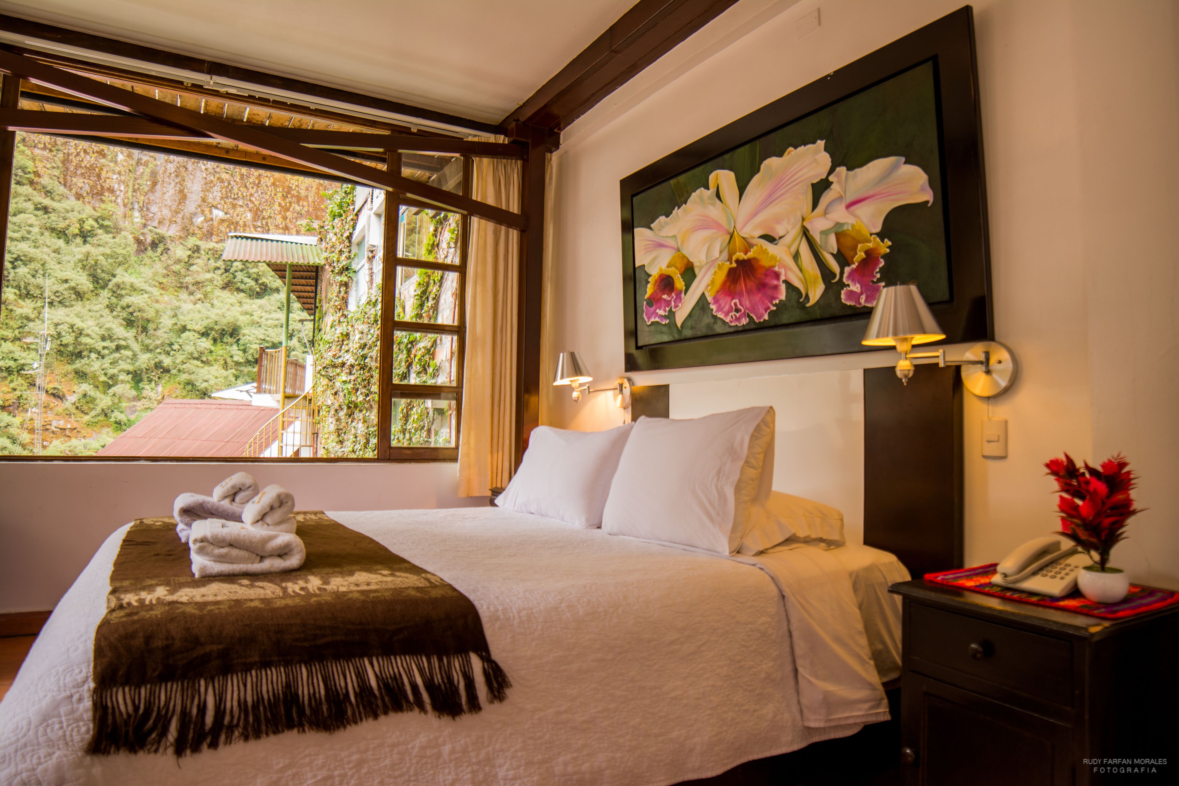 Una cama bien hecha con cobija y toallas, un cuadro de flores en la pared, una lámpara de noche y una mesita de noche con un teléfono y flores.