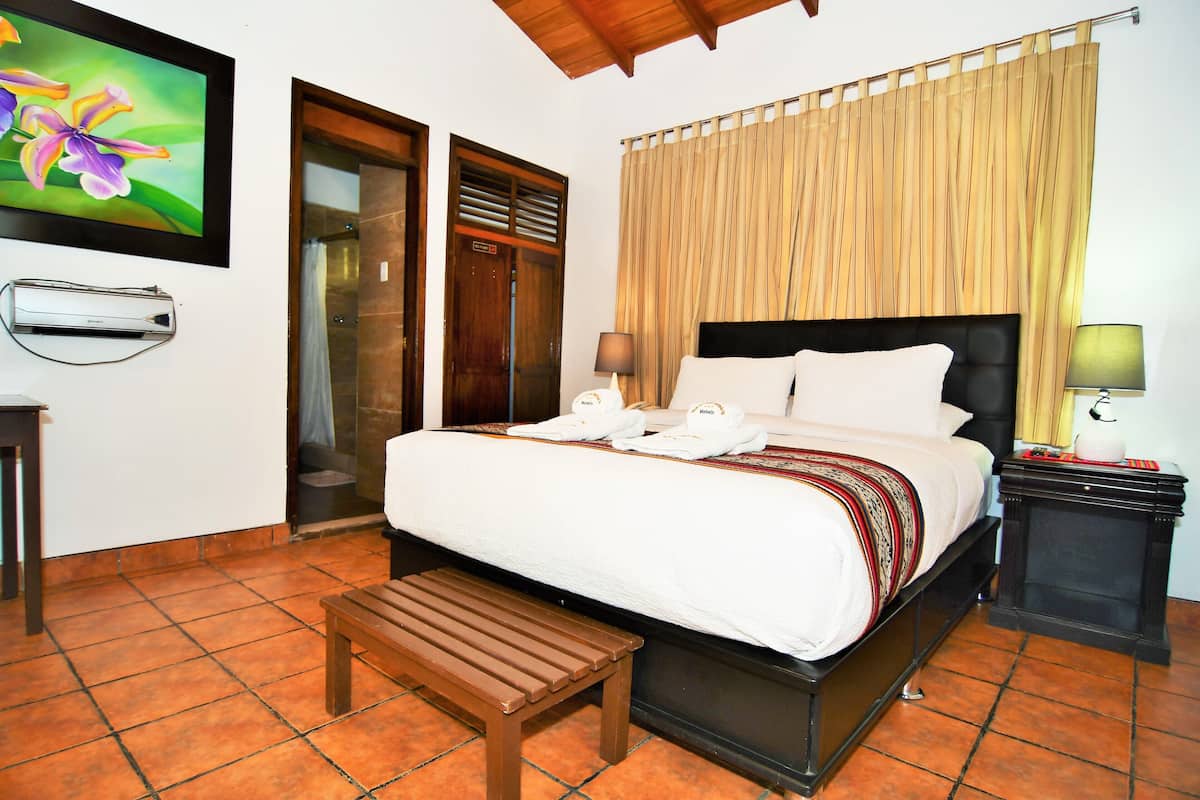 A hotel room with a bed, a bench, a bedside table, a lamp, and a wall-mounted air conditioner.