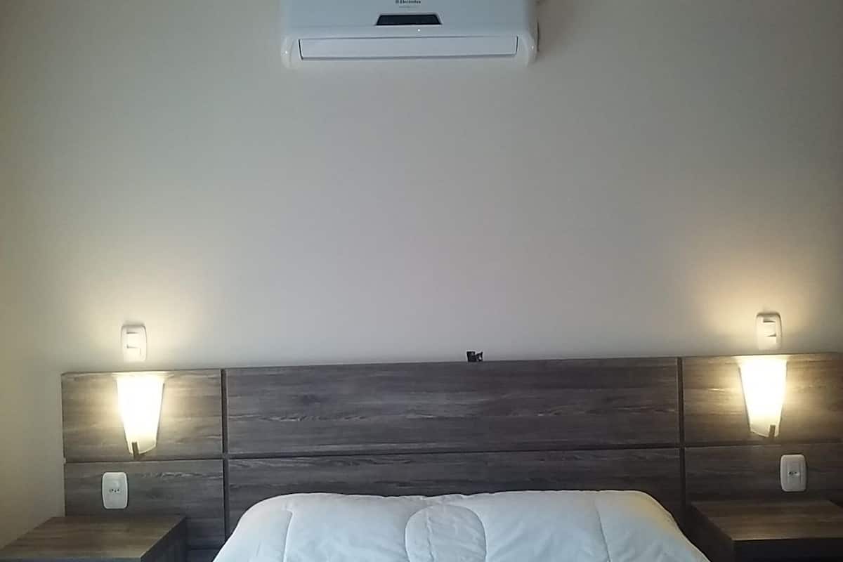 A hotel room with a bed, two bedside tables, a wall-mounted air conditioner, and two wall-mounted light fixtures.
