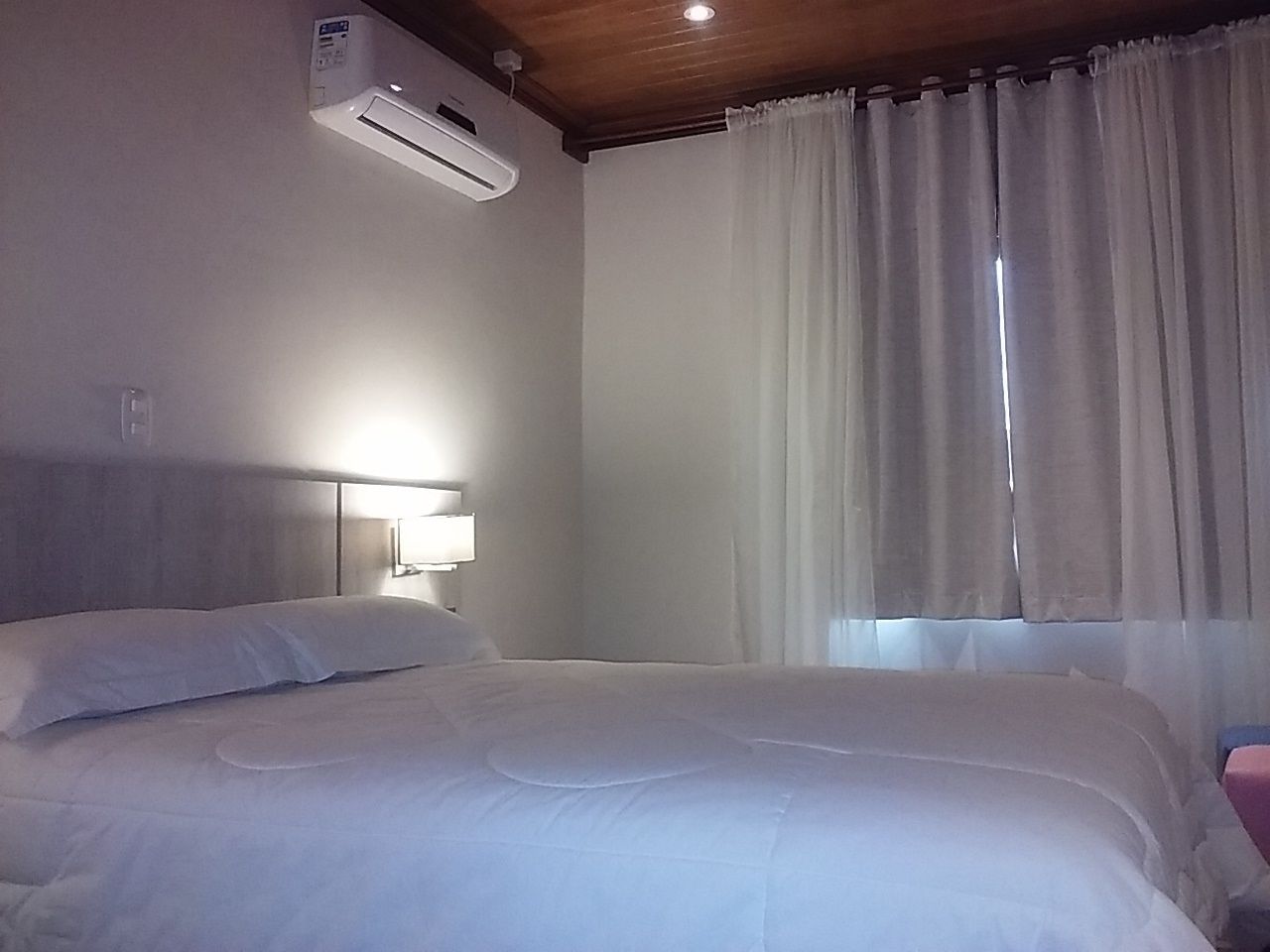 A hotel room with a bed, an air conditioning unit, a nightstand with a lamp, and a window with curtains.