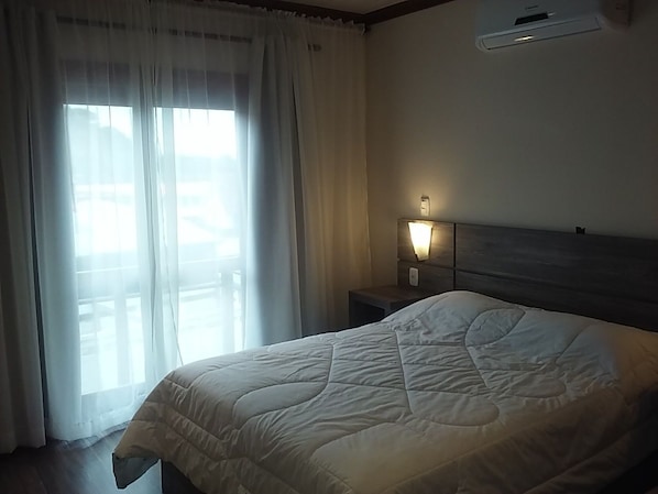 A hotel room with a bed, a nightstand, a wall-mounted air conditioner, and a window with sheer curtains.