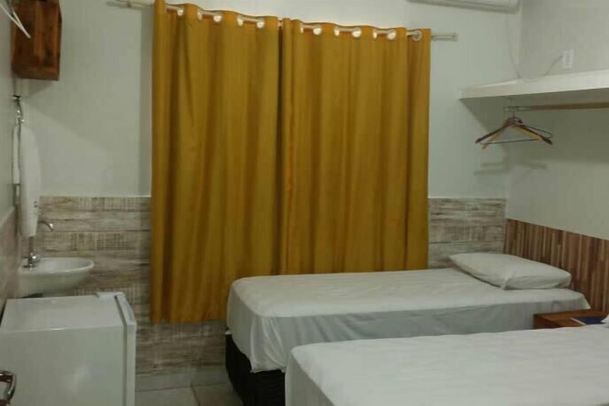 A small, single-room hotel room with a bed, a bedside table, a hanging clothes rack, and a bathroom with a sink and toilet.