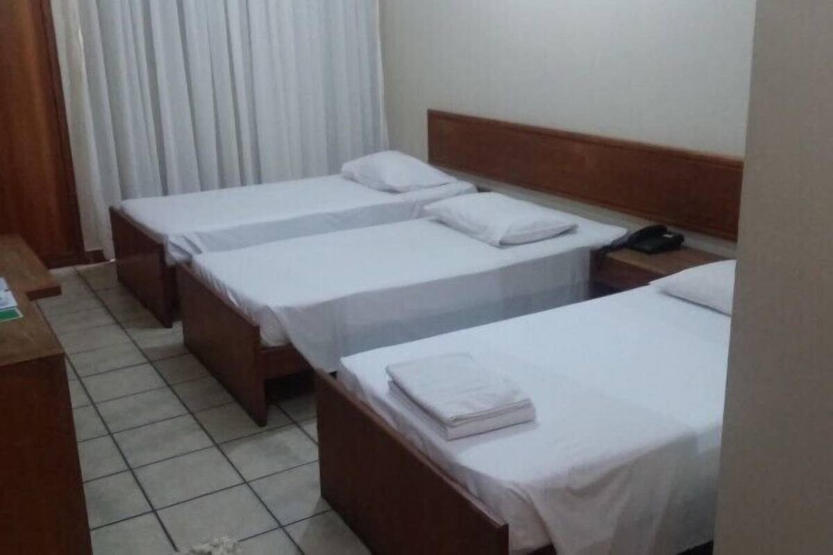 A hotel room with three single beds, each with a wooden headboard and white bedding. There is a desk with a telephone and some papers on it.