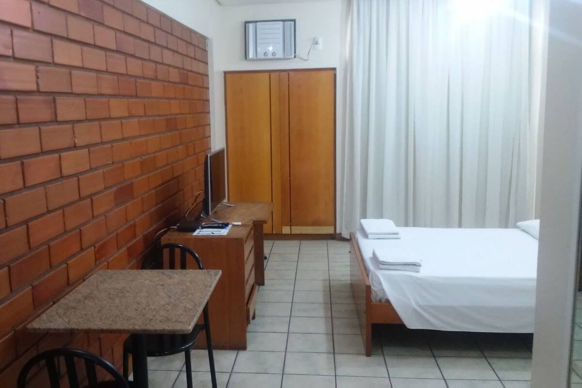 A small, compact room with a brick wall, a wooden desk with a chair, a bed with white bedding, and a wooden wardrobe.